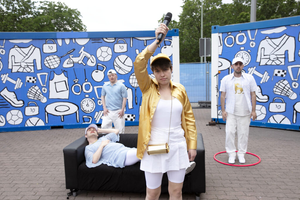 One person in the foreground looks into the camera and holds a microphone above him with one hand in the air. In the background there are more people in front of blue containers, one is lying on a couch.