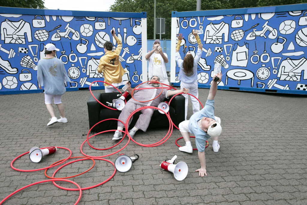A group of people are standing in a square. A person to the right in the foreground is doing a one-handed bridge and holding a water spray bottle in the air. Behind them, a person is lying on a couch, with many hula hoop rings on and in front of him. Around both of them are several megaphones on the floor. In the background people are dancing in front of two blue containers.