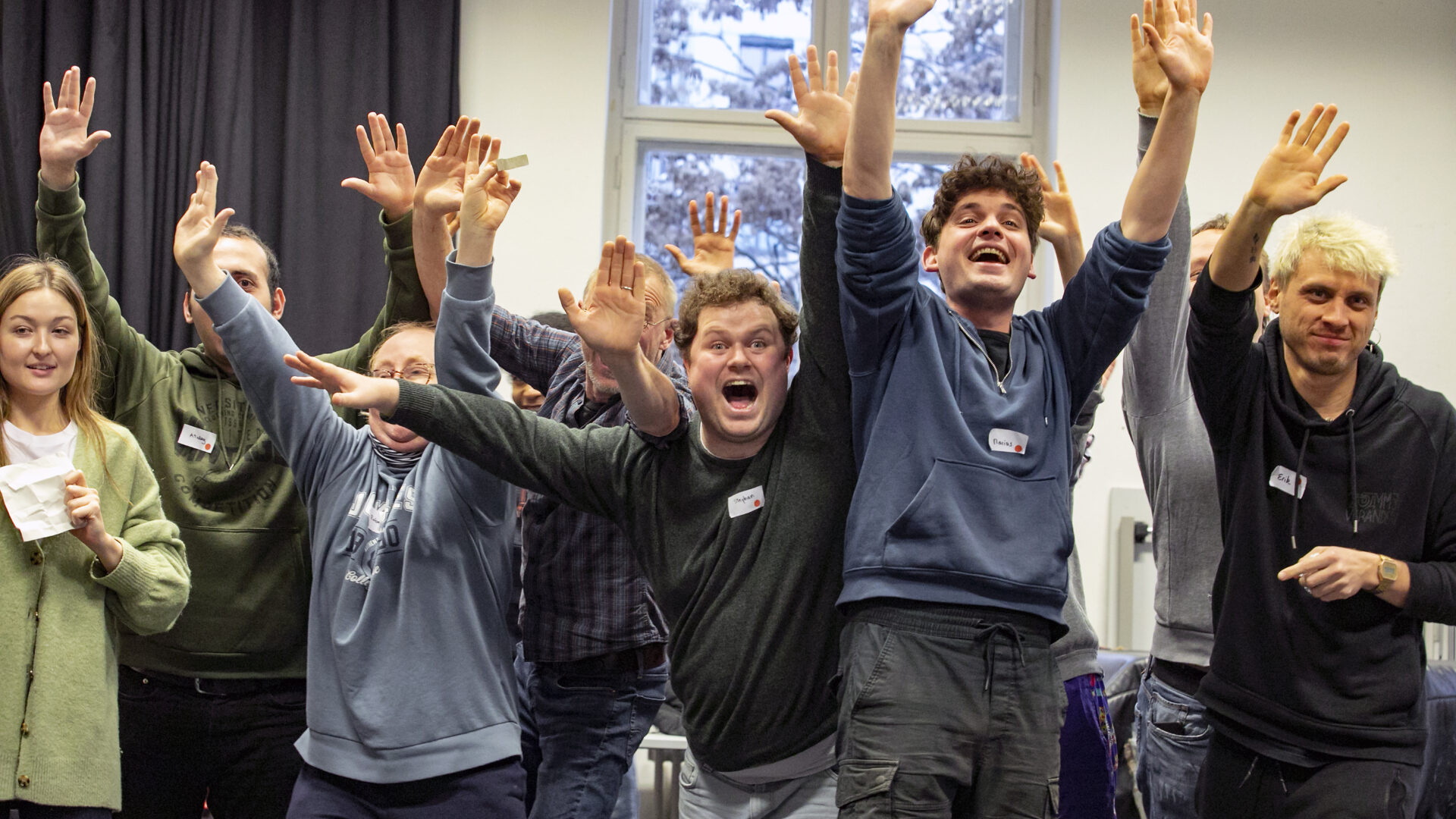 Eine Gruppe fröhlicher Menschen steht in einem Raum zusammen und hebt jubelnd die Arme, als würden sie gemeinsam feiern oder an einer lebhaften Gruppenaktivität teilnehmen.