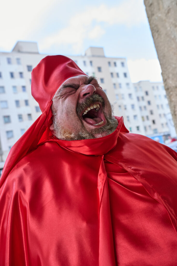 Ein Mann in einer roten, glänzenden Robe steht vor einem Hochhaus und schreit.