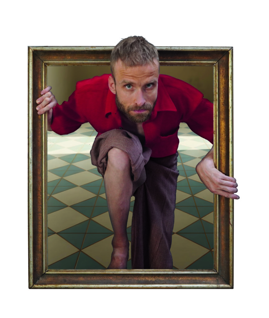 Ein Mann mit kurzem blondem Haar und rotem Hemd steigt scheinbar aus einem großen, aufrecht stehenden Bilderrahmen heraus. Er hat ein Knie nach vorne gesetzt und hält sich mit beiden Händen am Rahmen fest. Der Hintergrund zeigt einen geometrischen, gekachelten Boden. Die Szene wirkt wie eine optische Illusion, als würde der Mann aus dem Bild in den Raum treten.