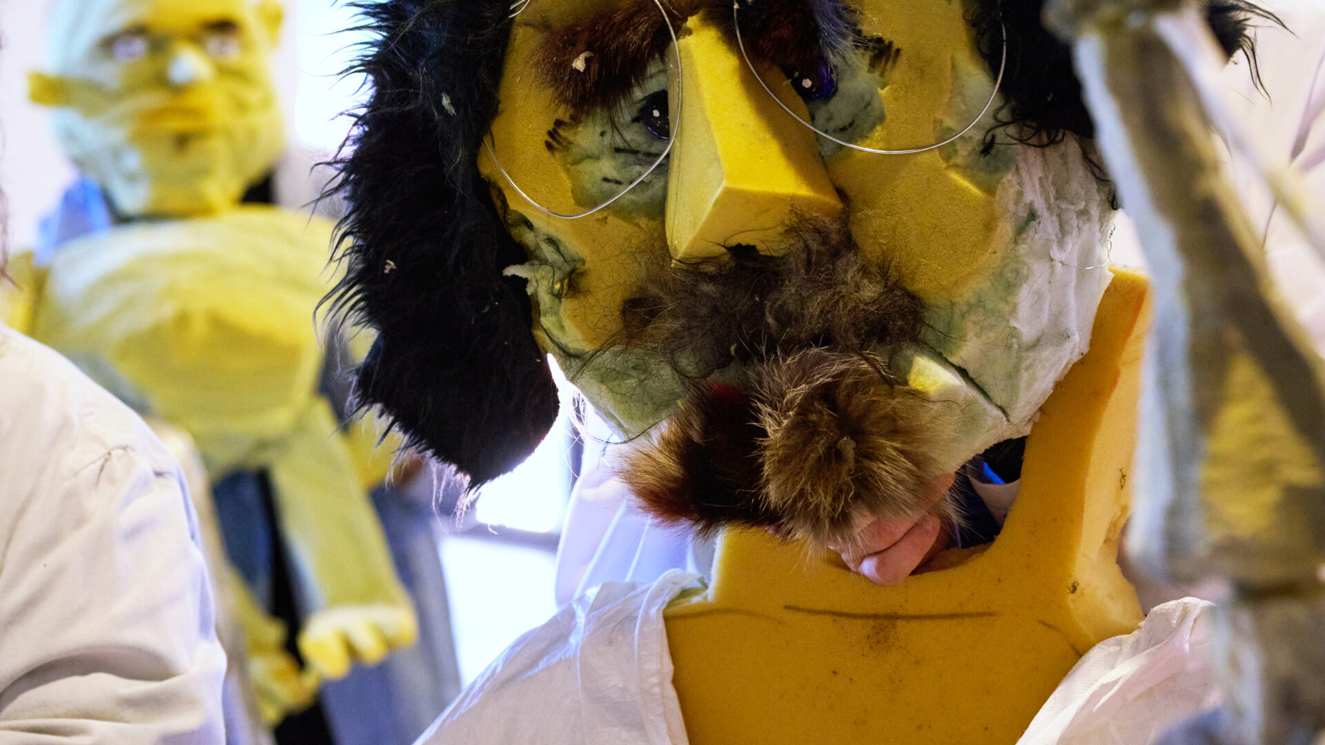 Eine große, skurrile Puppe mit einem gelben, grob geformten Gesicht aus Schaumstoff trägt eine schwarze Perücke, eine Brille und einen künstlichen Schnurrbart. Sie ist in ein weißes Gewand gehüllt und sitzt an einem Tisch mit weiteren Puppen im Hintergrund.