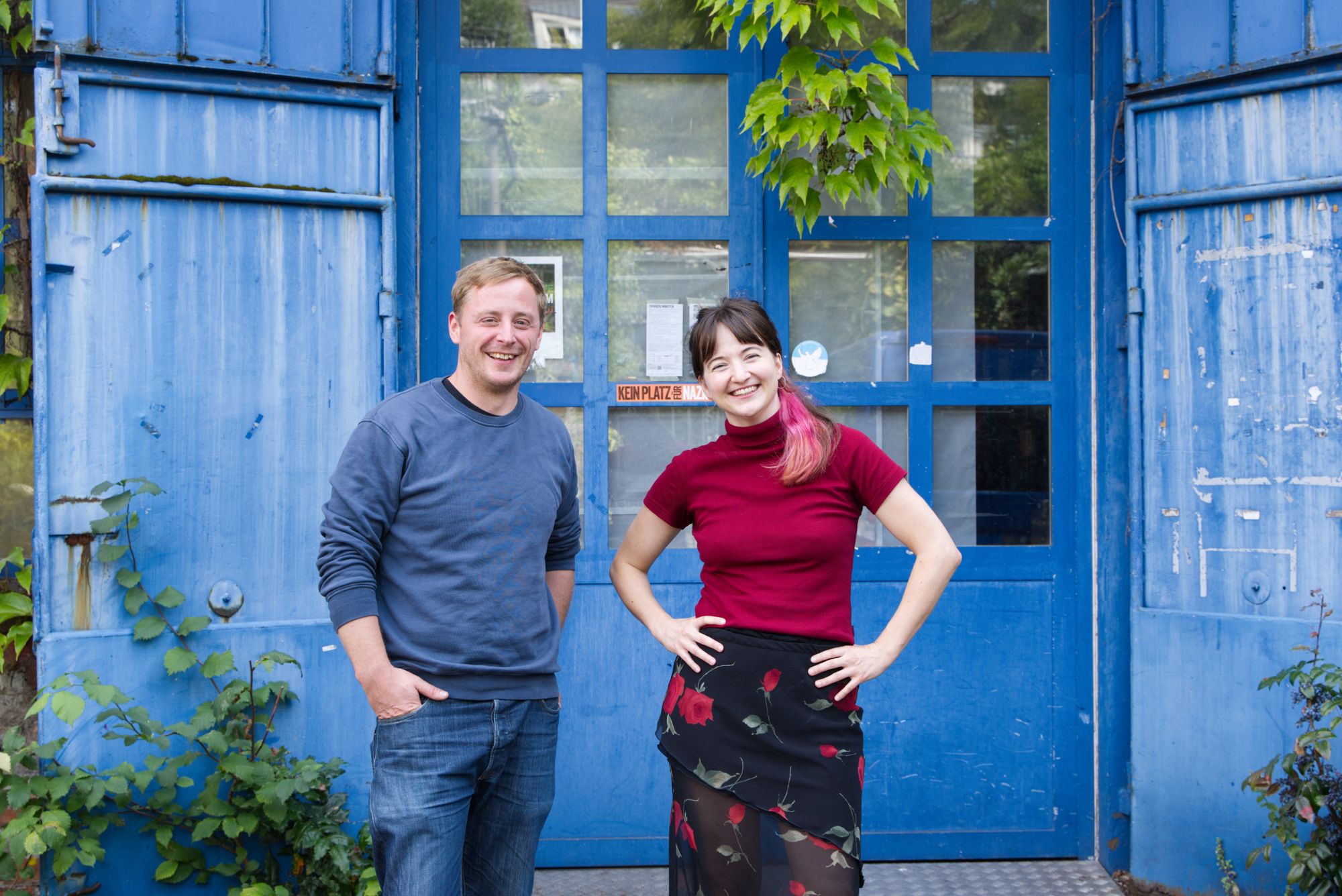 Ein Gruppenfoto vor der verwitterten blauen Metall- und Glastür. Mark Schröppel steht links in einem blauen Sweatshirt und Jeans, die Hände in den Taschen. Laura Besch steht rechts in einem roten Rollkragenpullover und einem Rock mit Rosenmuster, die Hände in die Hüften gestemmt. Beide lächeln. Über der Tür wächst Efeu, und links ist eine grüne Pflanze zu sehen. An der Tür ist ein orangefarbener Aufkleber mit dem Text „KEIN PLATZ FÜR NAZIS“ sichtbar.